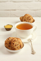 Oatmeal cookies with walnuts and raisins on small saucers and a cup of green tea, standing on a white wooden table against a white brick wall.