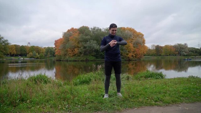 Amateur stout athlete in sport outfit doing exercises in the park near pond with autumn trees background before the run. Healthy lifestyle motivation concept.