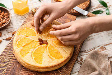 Woman decorating tasty orange pie with almond nuts on light wooden background