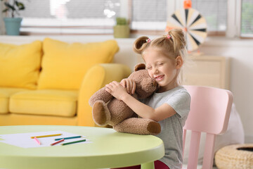 Cute little girl with ponytails hugging teddy bear at table in living room