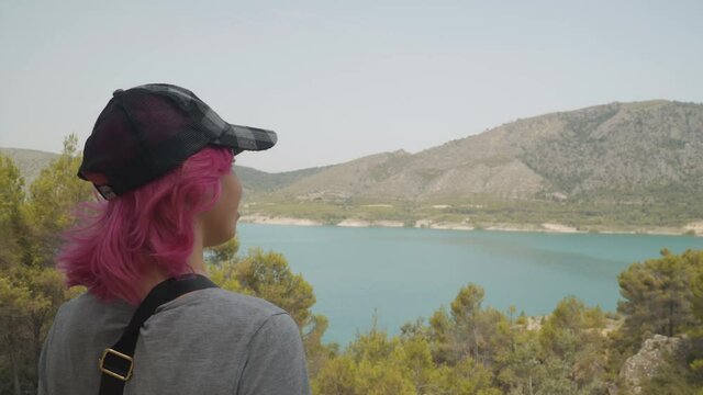 Mujer de pelo rosa y gorra  mirando el embalse desde lo alto de la colina, mujer de pelo corto mirando el lago de agua turquesa, mujer feliz  mirando las monta&ntilde;as y el agua , mujer haciendo senderismo