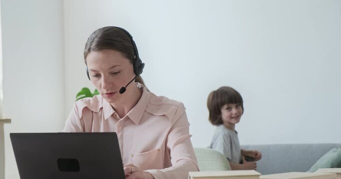 Young female tutor wearing headset teaching students online in video conference. Young cute child 6 years old distracts her mother from remote work.