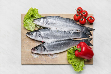 Board with fresh uncooked sea bass fish and vegetables on white marble background