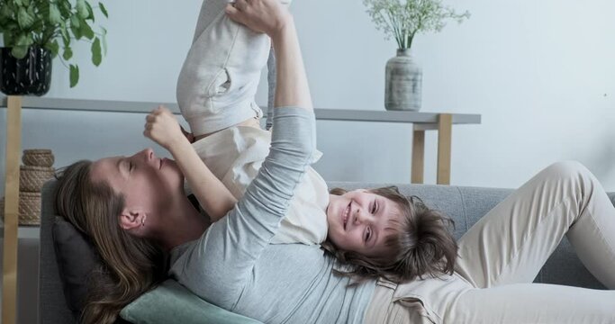 Carefree joyful cheerful boy son bouncing fun playing and cuddling young mother lying on the couch. Relaxed family day of loving parents and children
