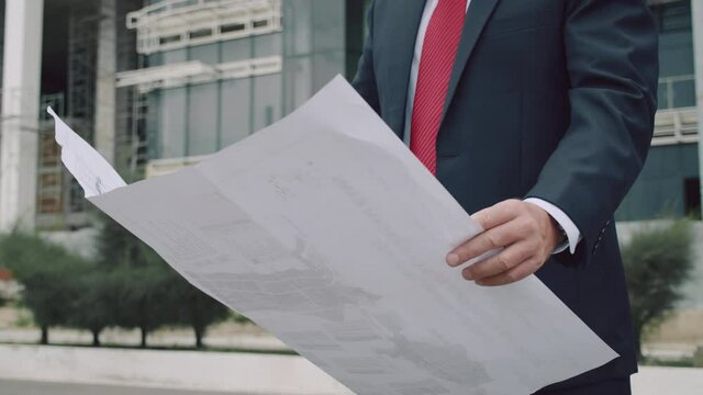Engineer helmet checks a construction plan in new microdistrict of the city.Senior Engineer Planning Manufacture Work business suit a red jacket with construction card in his hands