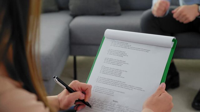 Close up over the shoulder woman psychologist taking notes on clipboard and listens patient at psychotherapy session. Unfocused man with mental health problems sitting on couch talking to therapist