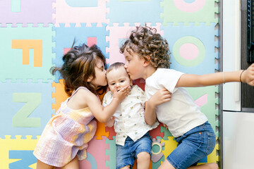 Top view of little girl and boy kissing to baby brother lying on floor in playroom