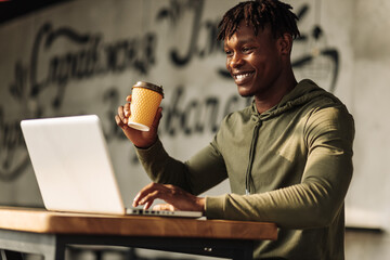 African American man drinks coffee or tea from a paper cup, prints at the table on a laptop. work at table with a laptop