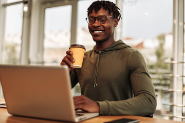 African American man drinks coffee or tea from a paper cup, prints at the table on a laptop. work at table with a laptop