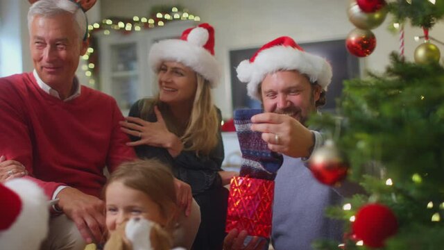 Father unwrapping present of socks as multi-generation family exchange gifts around christmas tree at home - shot in slow motion
