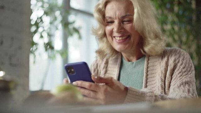 A woman aged sitting at a table and watching a video on the phone. She smiles sweetly and laughs