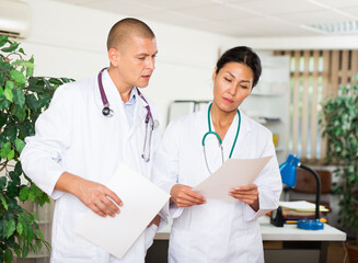 Fototapeta premium Young american doctor and asian nurse working together with case histories in modern clinic