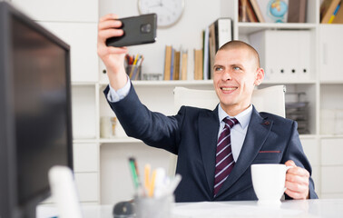 Business consultant male working in an office takes a selfie while sitting at the workplace with a cup of coffee