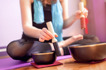 Low section of a young caucasian woman playing tibetan singing bowls over a purple yoga mat on wooden floor. Concept of meditation and relaxation at home. Yogini. Selective focus