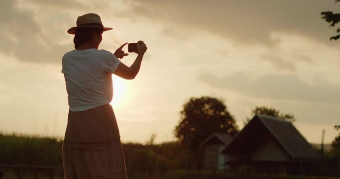 A female tourist in a brown hat, white T-shirt and brown skirt standing on the meadow use her mobile phone to take pictures of the sunset, beautiful orange flare on the farmer village at countryside. 