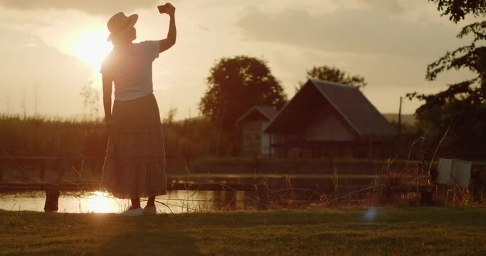 A female tourist in a brown hat, white T-shirt and brown skirt standing on the meadow use her mobile phone to take pictures of the sunset, beautiful orange flare on the farmer village at countryside.