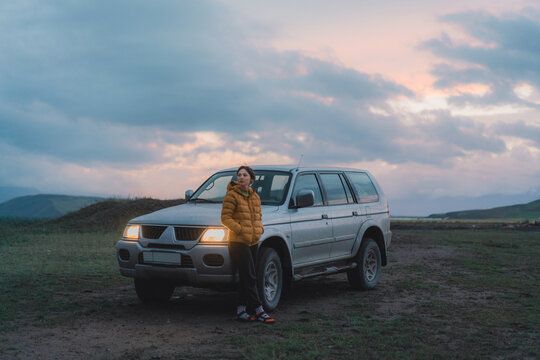 Woman standing near SUV car on dirt road at sunset 