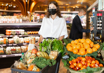 Young woman in protective face mask standing with full shopping cart while shopping in greengrocery during coronavirus pandemic