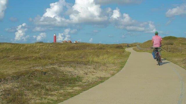 Slow motion of a bicyle path in the dunes. people riding bicycles through the beuautifull landscape. In the distance we see a lighthouse in the dunes. The sky is blue, clouds passing by. Serie of 7