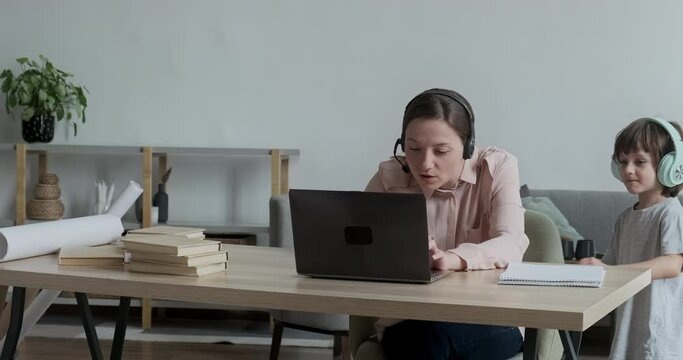 Young woman wearing headset working remotely from home on a video call, little son runs up and distracts and disturbs mom. Difficulties of online work and learning with young children.