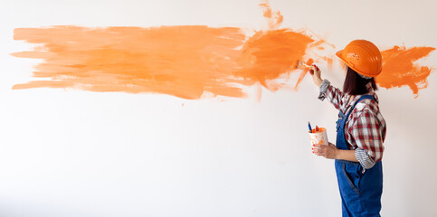 A woman in overalls and a helmet paints the wall in the room with orange paint with a brush. The concept of home renovation and painting the walls.