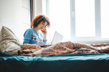 Curly woman talking on the phone straight from the bed noting homework. Online lessons