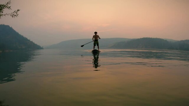 Man on Paddle Board Passing by Camera on Calm Water with Hazy Sunset