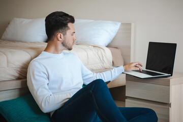 Fototapeta premium Man sitting on the floor leaning on the bed and typing on the computer. Online career. Internet technology. Sitting man. Computer technology. Business