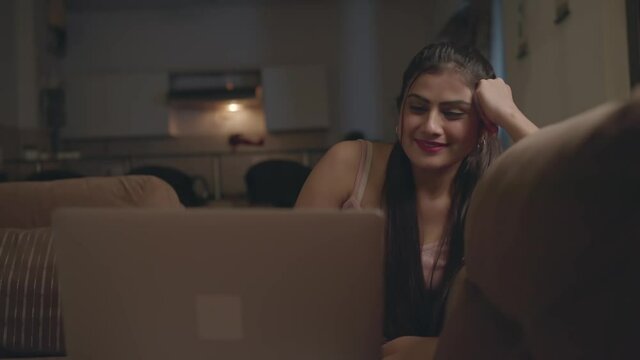 A young smiling modern Indian woman or female watching a movie or web series online or on OTT platform on a laptop in an interior house or home in the night