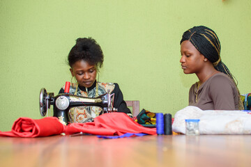 A female African fashion designer or tailor teaching her apprentice how to make clothe using sewing...