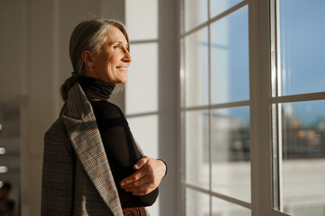 European senior woman with grey hair smiling while looking out window