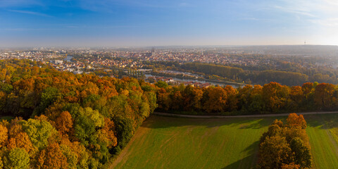 Drone point of view over skyline of Regensburg, Bavaria behind beautiful colorful variegated autumn foliage of deciduous forest on sunny day in October