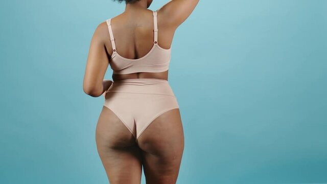 Rear-view medium portrait of curvy young African-American woman in underwear dancing tenderly on isolated blue background. Concept of natural beauty and body positivity