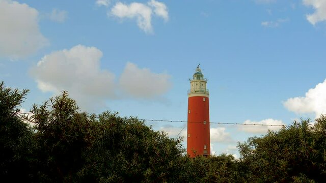 Time lapse wide angle steady shot of sea ​​buckthorn in the foreground. In the background a lighthouse in the dunes. The sky is blue with beautiful clouds passing by.