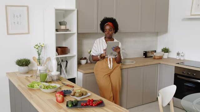 Medium slowmo shot of young African-American woman in casualwear using digital tablet while standing at bright modern kitchen with fresh fruit and vegetables on wooden table