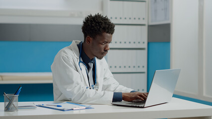 Obraz premium Young person working as doctor in medical office with laptop and document files. Medic wearing white coat and stethoscope sitting at desk using technology and tools in hospital room