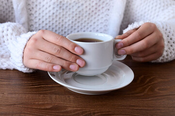 girl's hands in warm knitted white sweater holding cup of tea on wooden table