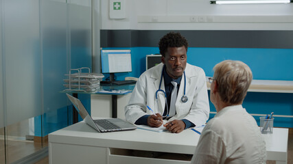 Doctor with healthcare expertise consulting elder woman sitting at desk in medical office. Young medic in uniform giving advice to sick old patient at hospital facility. People discussing disease