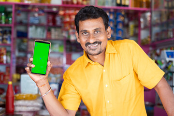 Smiling Merchant at groceries store hold mobile with green screen mock up by looking at camera -...