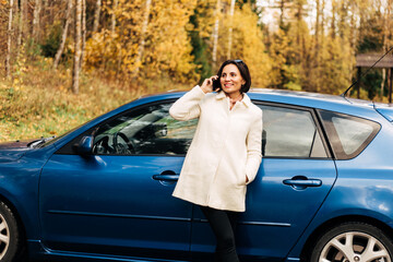 Fashion business woman in white female coat calling on cell phone outside her car in autumn park