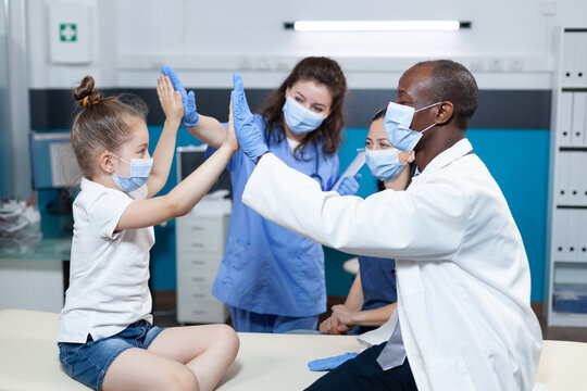 Girl Patient With Medical Protective Face Mask Against Coronavirus Giving High Five To Medical Team During Clinical Appointment In Hospital Office. Pediatric Doctor Discussing Healthcare Treatment