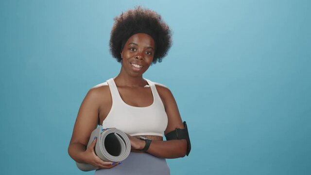 Medium studio portrait of smiling young curvy African-American woman in tight sportswear and armband for smartphone posing for camera with yoga mat on blue background