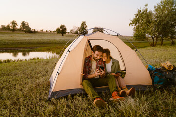 White couple using cellphones and sitting in tent during camping - obrazy, fototapety, plakaty
