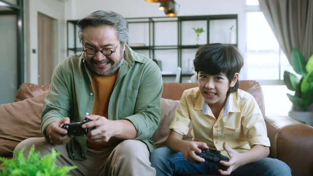happiness cheerful asian dad son play video game together at
 home fun and excited hand hold joystick in quarantinem oment entertainment at home concept.Smiling family sitting on the couch together