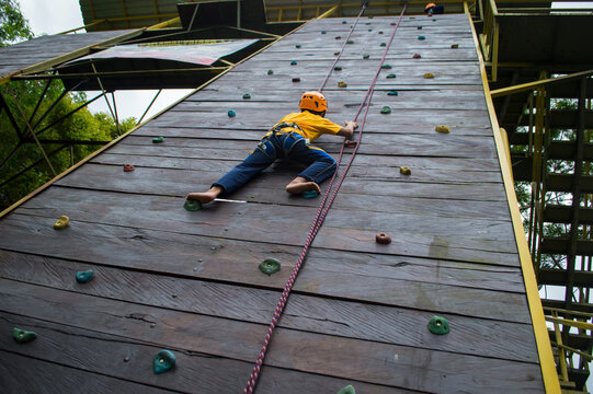 A nine-year-old boy equipped with a rock-climbing gear kit is climbing a wall outdoor with confidence (low angle)