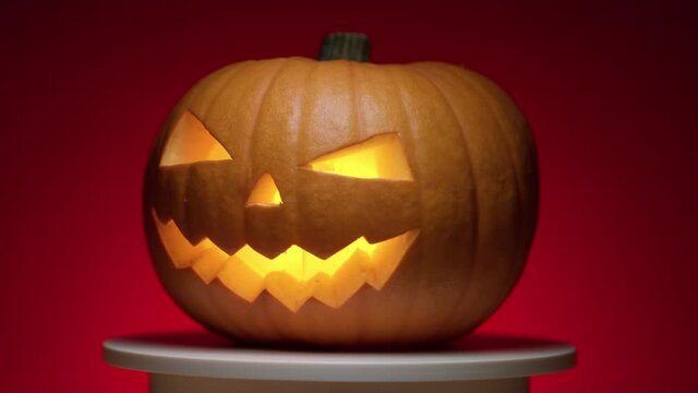 continuous looping Rotation of a Halloween pumpkin with carved teeth, eyes and nose, with a glowing fanar inside on a red background with a transition to darkness
