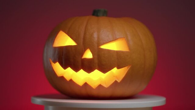 continuous looping rotation of a halloween pumpkin with carved teeth, eyes and nose, with a glowing fanar inside on a red background with a smooth fade into darkness