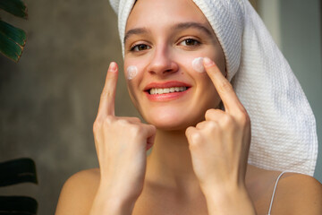 Close-up shot of a beautiful young woman doing her morning routine, applying face cream while covering her hair with a towel after washing.