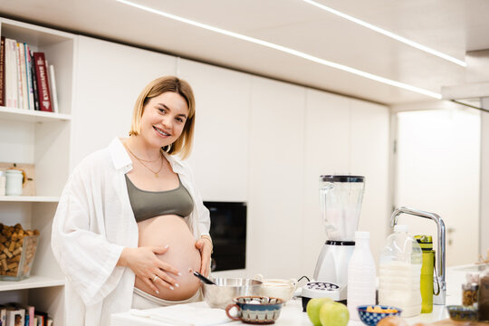 White pregnant woman smiling and touching her belly while cooking