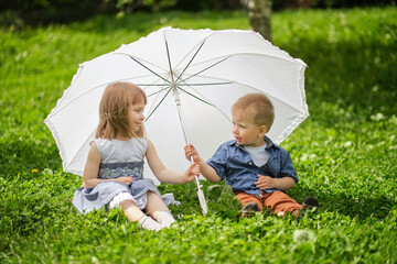 little girl and boy are hiding under an umbrella. brother and sister are playing in the park.